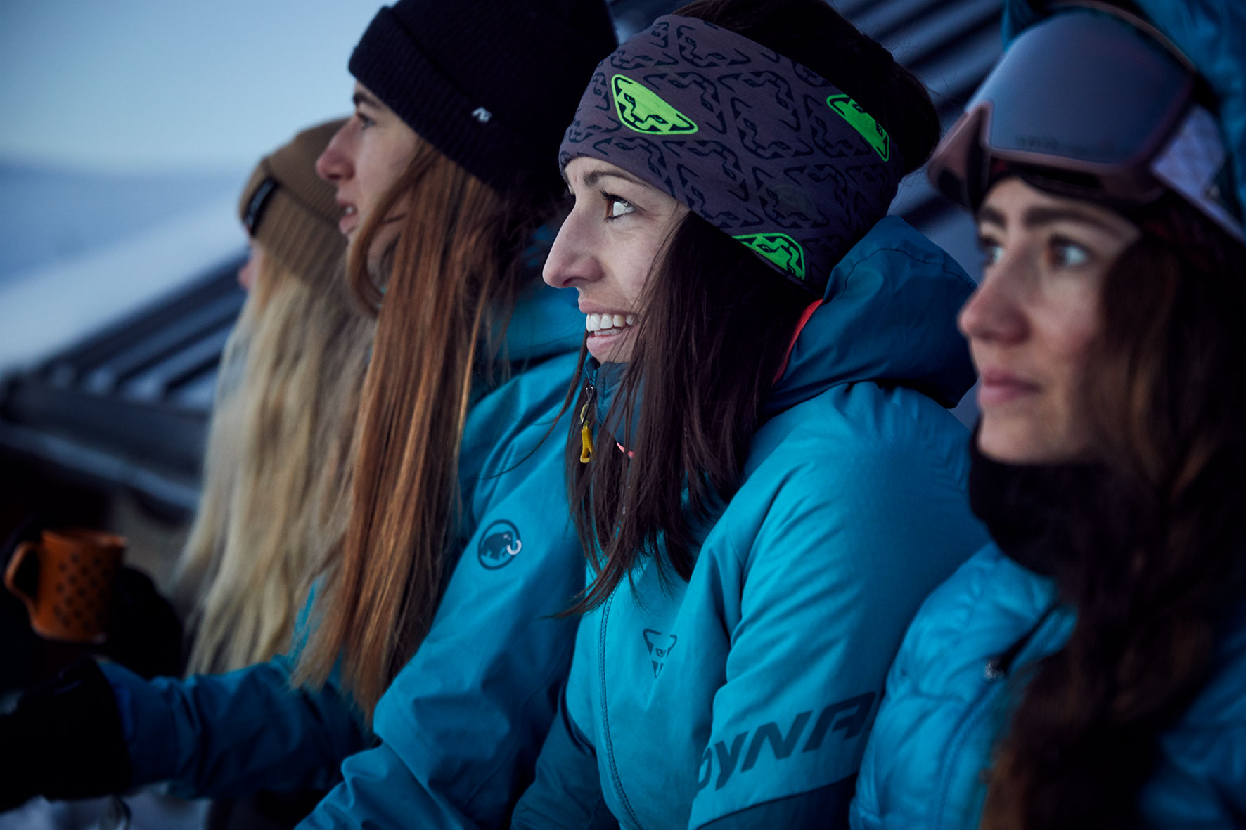 Four women in outdoor sport sitting against a night hut watching the sunrise over the mountain, dressed in ski clothes ready to go on a ski tour.