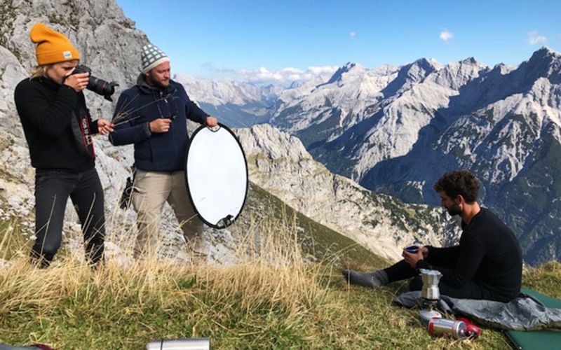 Professional photographer Jessica Zumpfe standing on top of the Karwendel mountain range while shooting a sports model for hess natur sustainable fashion brand. Next to her, her lighting assistant with a lighting reflector.