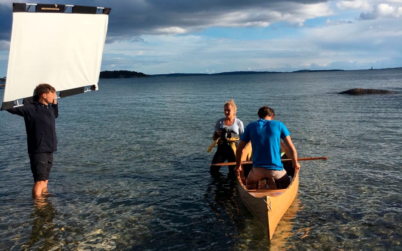 Professional Photographer Jessica Zumpfe standing in the sea in her wetsuit and shooting a sports model in a wooden canoe. Her assistant is standing next to her with a light reflector.