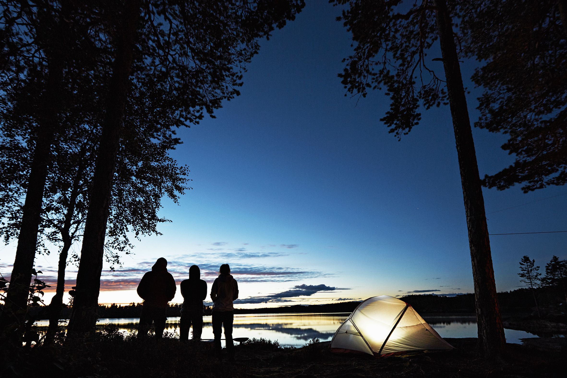 Silhouette of three people standing next to their lit tent under black trees by a lake near Oslo, Norway.