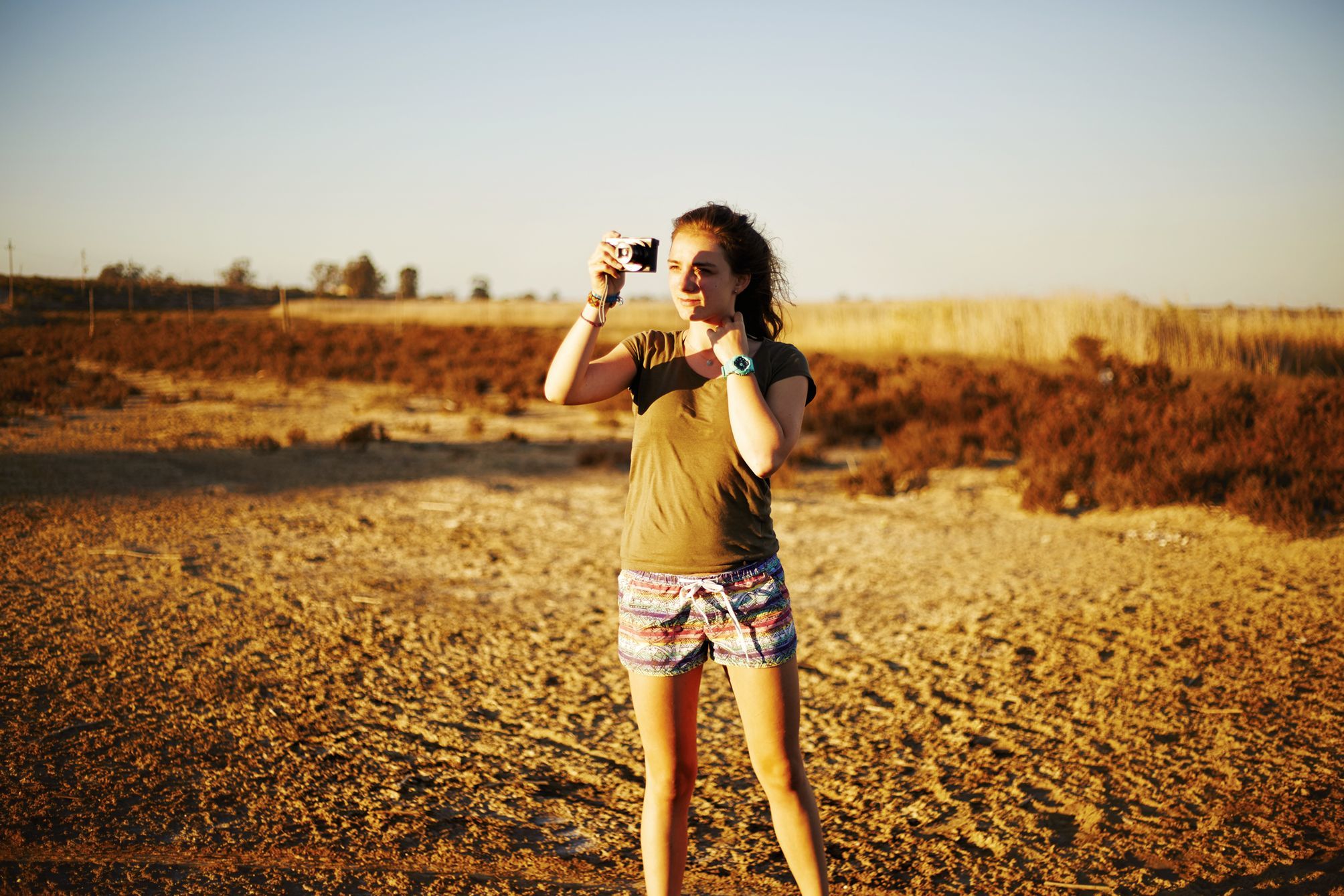 Young girl standing in the middle of the desert holding a camera to her face and taking a picture.