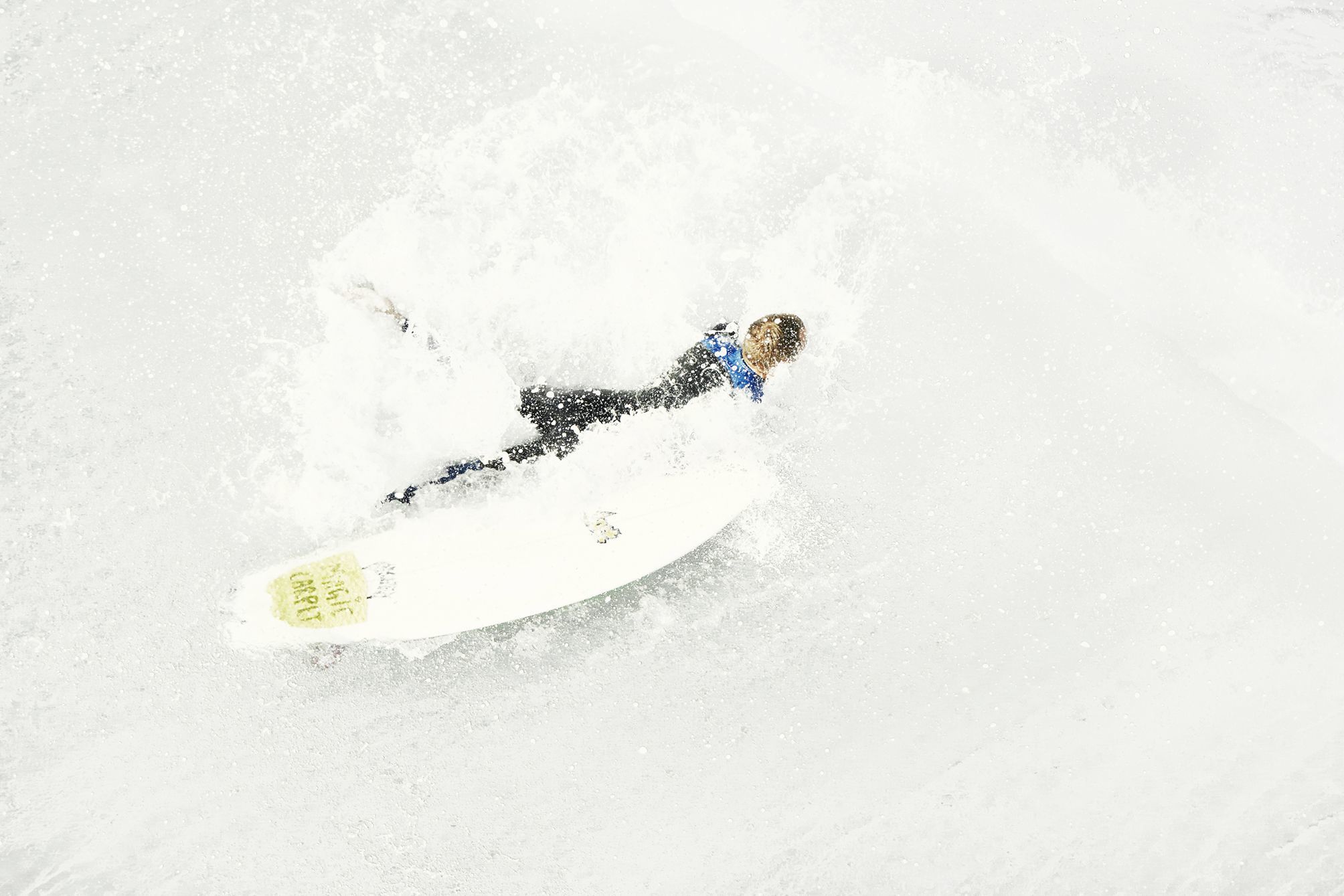 Surfer shot from above crashing next to his surfboard with a magic carpet sticker as a pad.