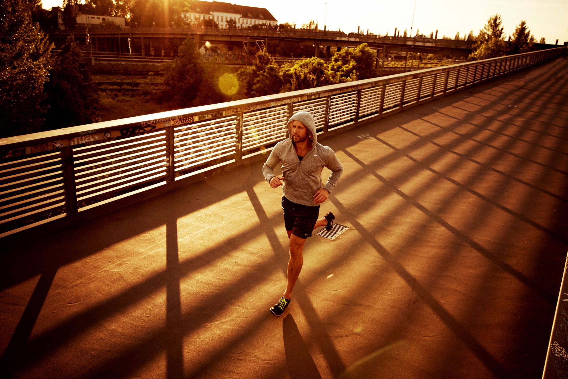 Urban sports trainer running over a bridge in the north of Berlin, the sun casting shadows from the reilings.