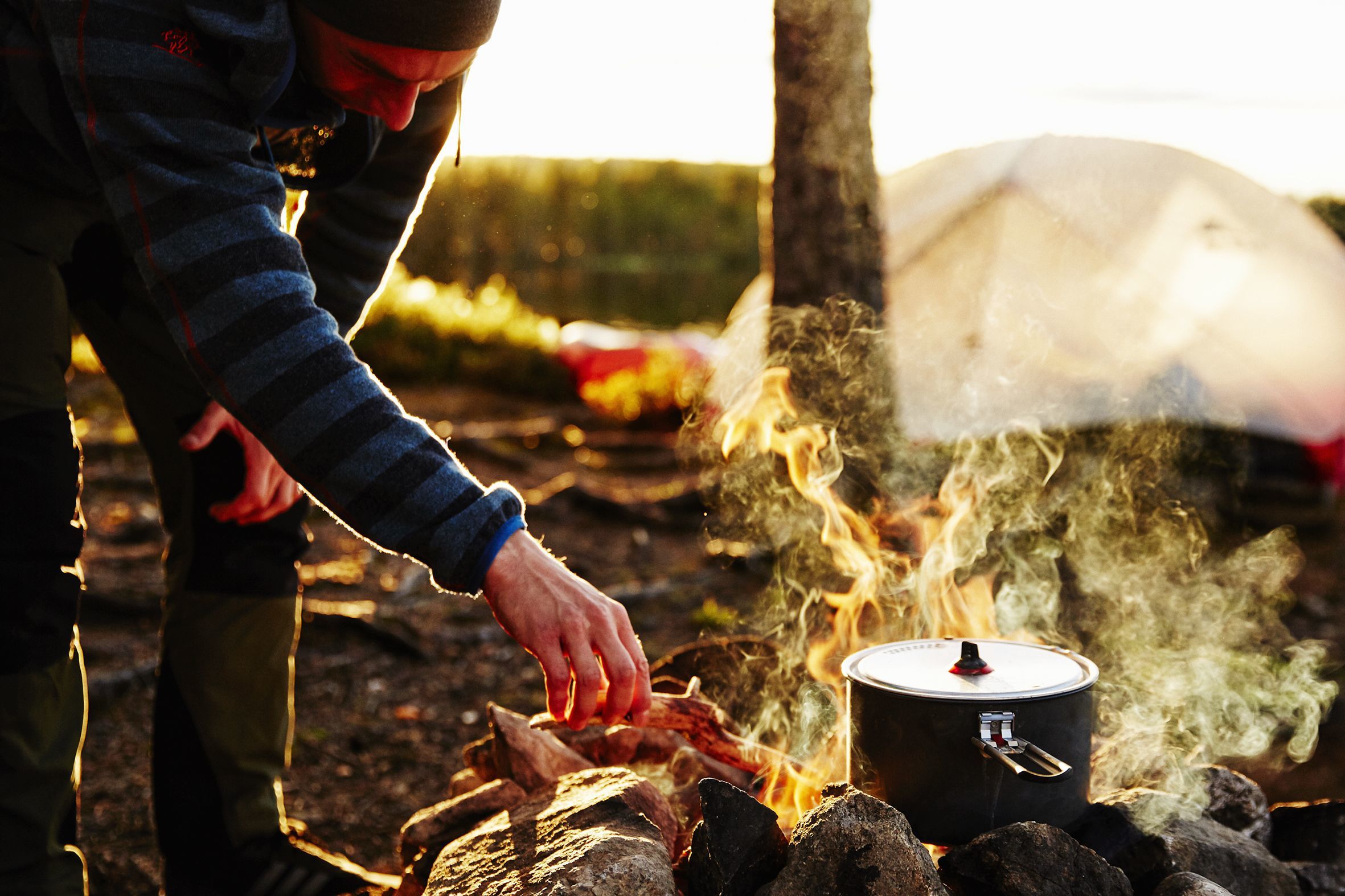Young man stoking the fire at a campite of our expedition in norway, while cooking food over the fire.