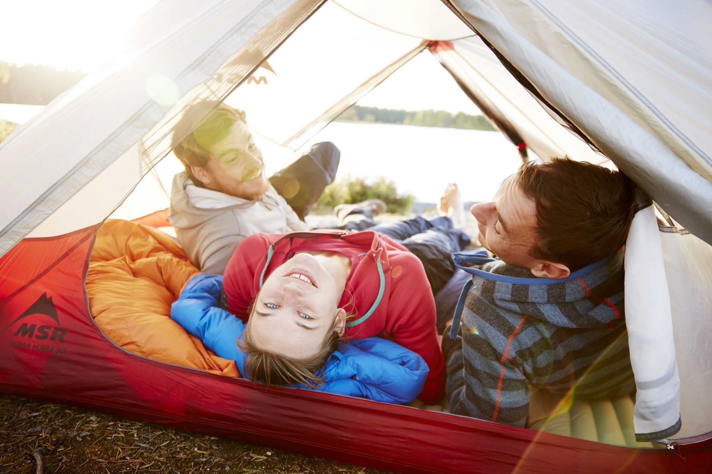 Group of friends lying in a tent laughing and looking up at the viewer. In the background is the lake district of Oslo, Norway