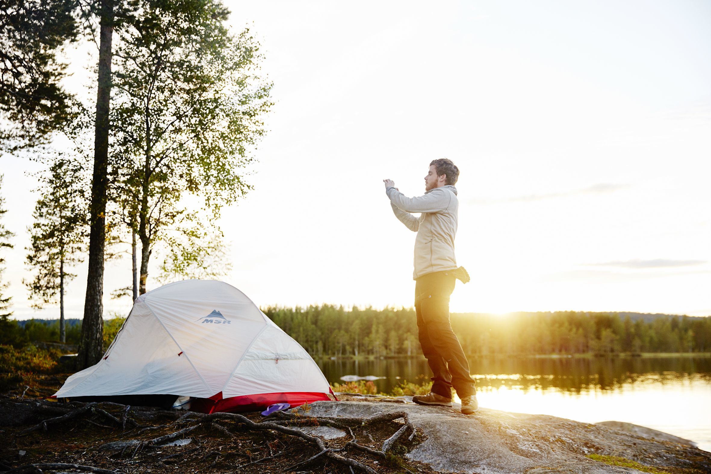 Young man taking a picture of his tent on his cellphone at sunrise near a lake in Olso, Norway.