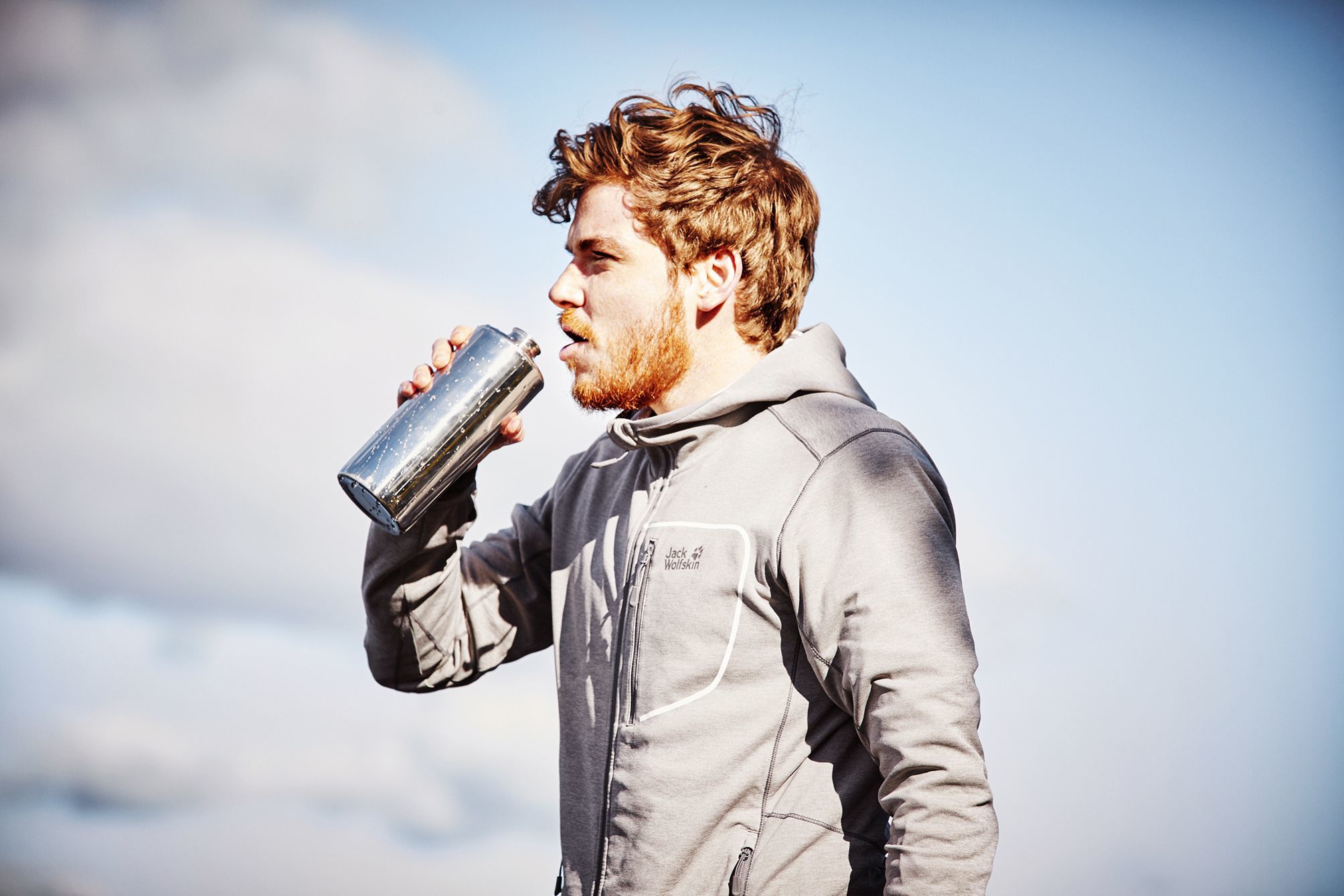 Young man juxtaposed infront of a semi cloudy sky in his jack wolfskin jacket drinking from a