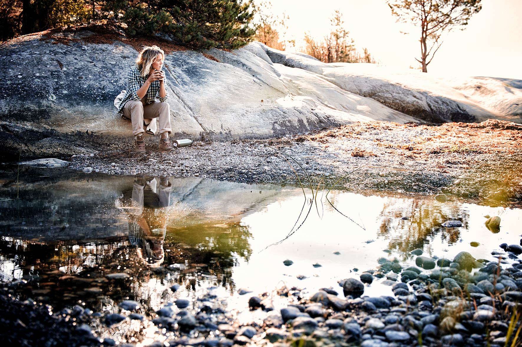 Hannah sitting on a rock at sunrise drinking coffee, being reflected in the puddle in front of her. She is wearing fair fasion from Hess Natur.
