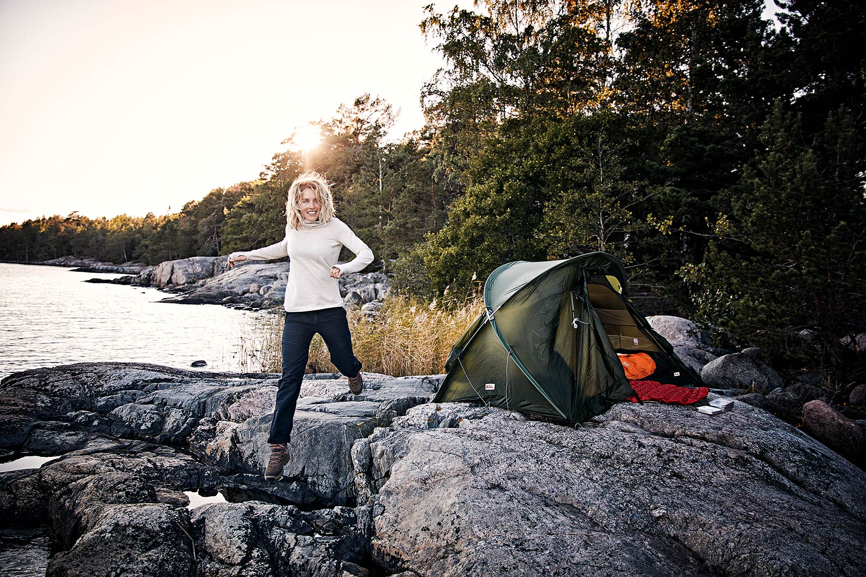 Fair fashion model Hannah, jumping over the rocks in front of her tent by the sea at sunrise in Stockholm, Sweden