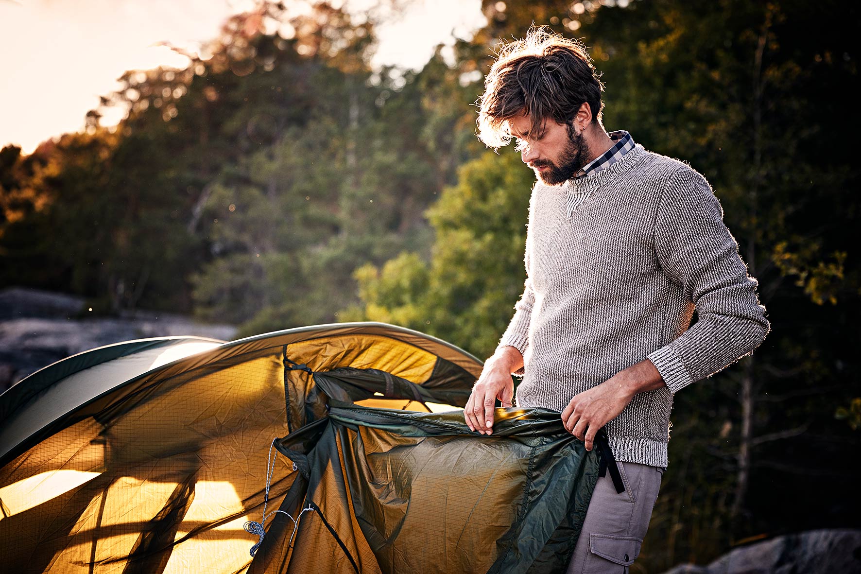 Young man rolling up his tent at dawn while wearing fair fashion clothes near Stockholm, Sweden.