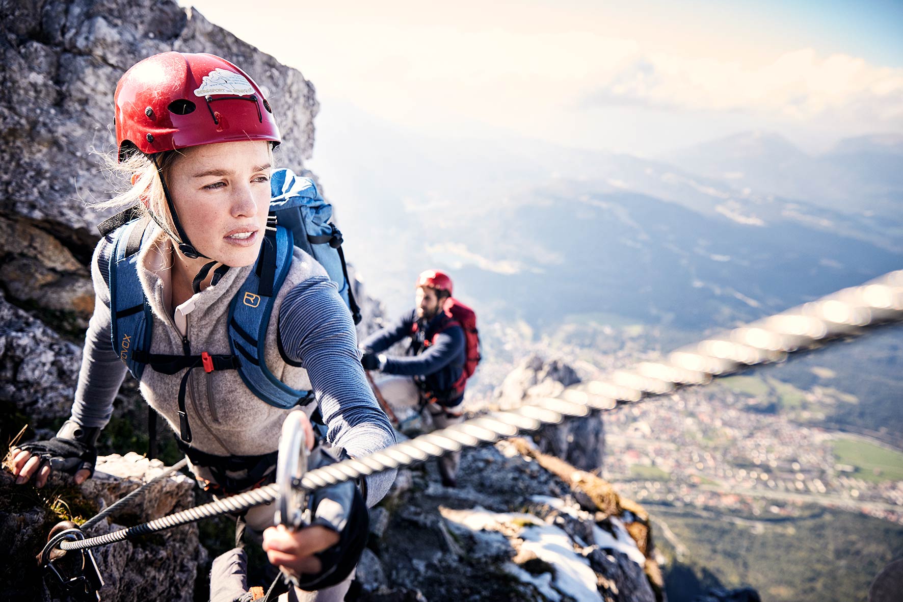 Sports photography models Hannah and Carlo climbing a via Ferrata at the Karwendel Mountain range, with Mittenwald in the background and far below them.