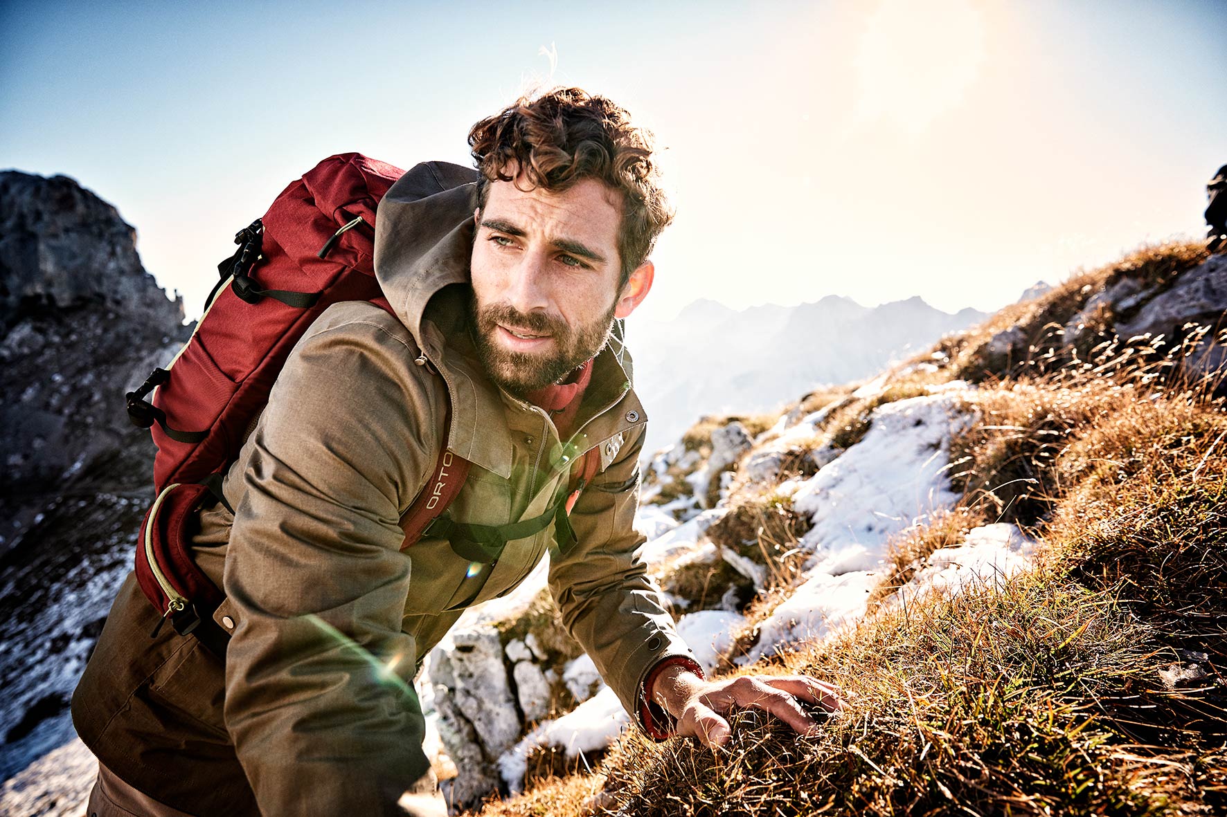 Young sports model carrying his ortovox backpack up the mountain in spring, with a few snow fields behind him.