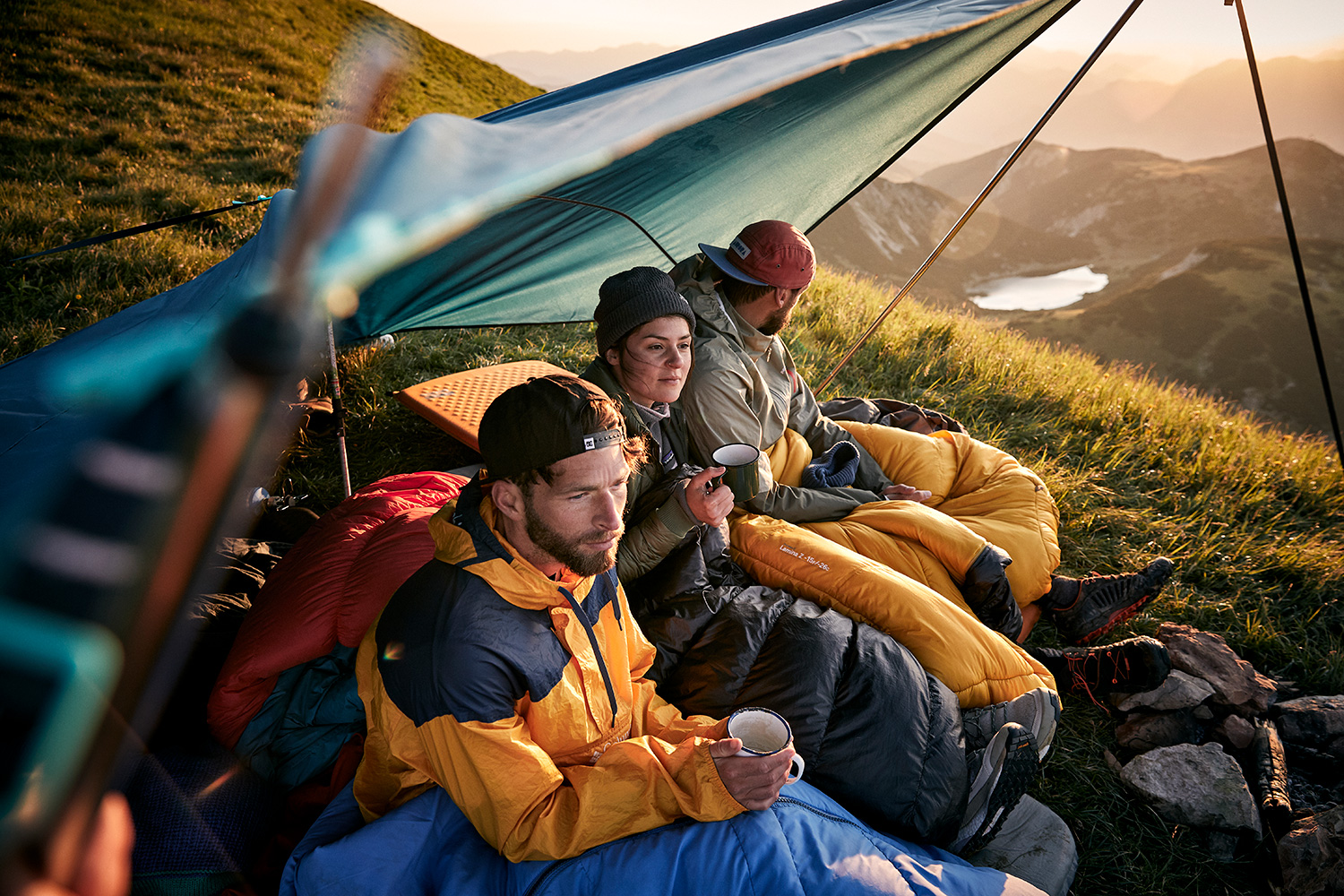 Three friends drinking coffee in the sunrise under a tarp ontop of the Rofanspitze in Austria