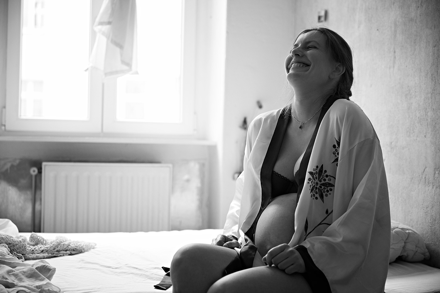 Black and white portrait of pregnant johanna sitting on a bed and laughing