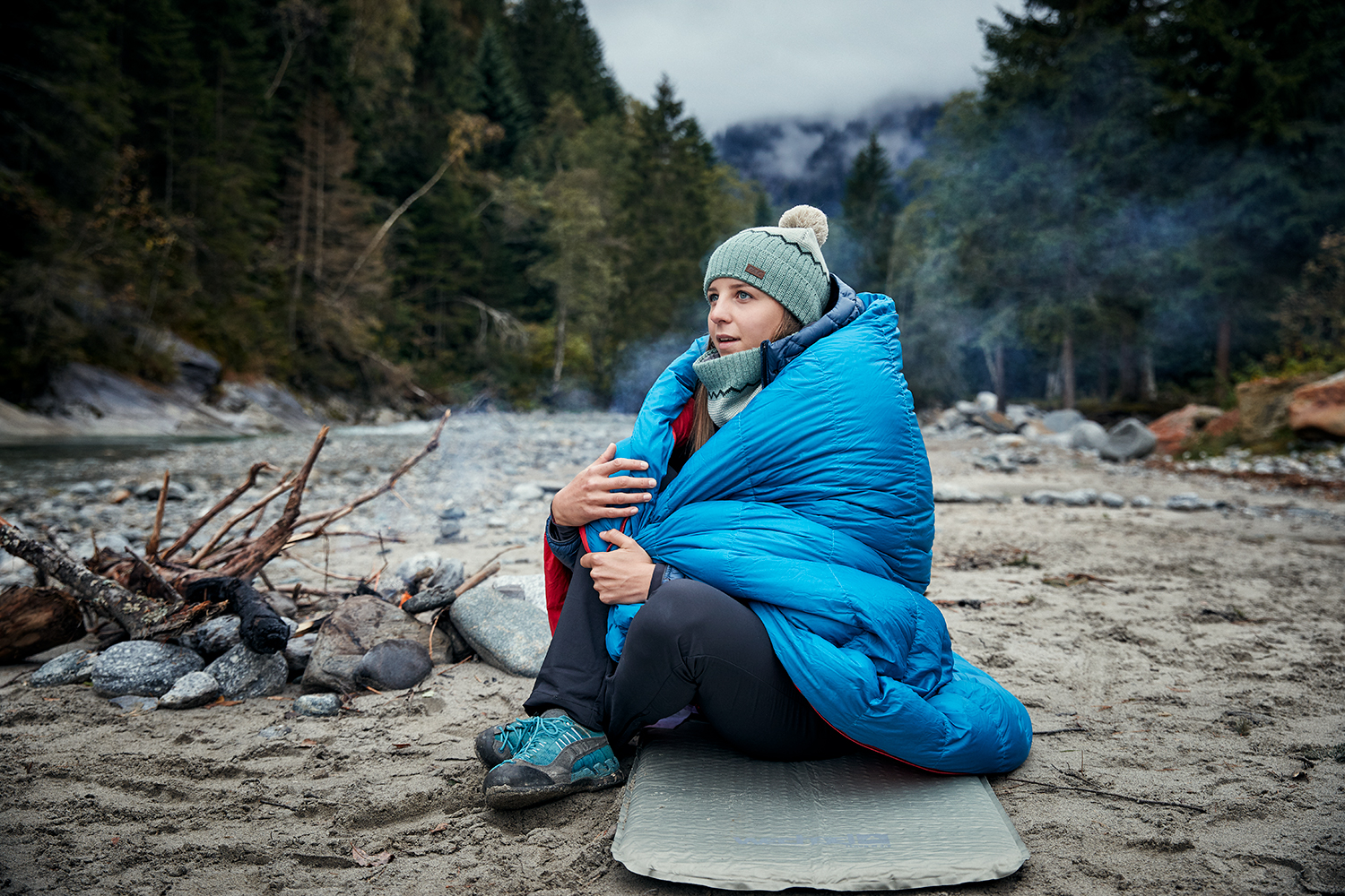 Woman sitting by a fire next to a river bed, cuddled in her sleeping bag by a river in Ginzling