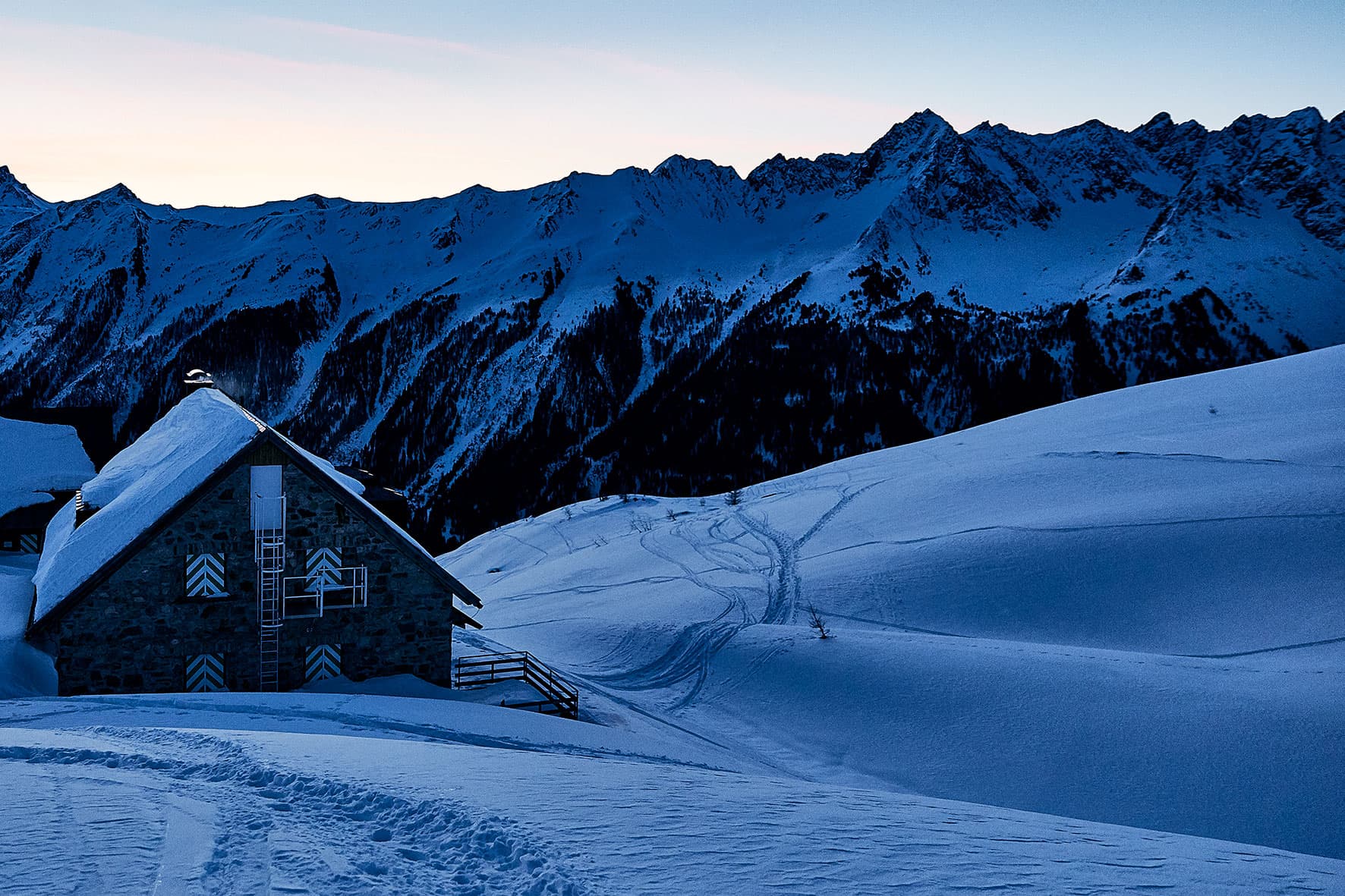 Photo of a winter night shelter ontop of the mountain before sunrise, the ski lines leading away from the hut and down the hill, with the mountain black in the background.e