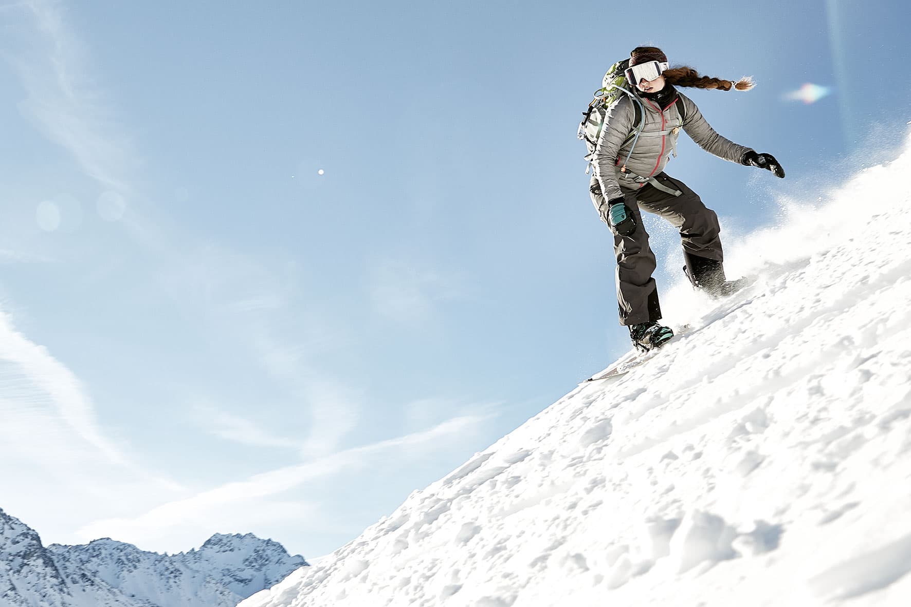 Anja showing what women in outdoor sport can do, snowboarding down a steep slope with the spray behind her and the mountain in the background.